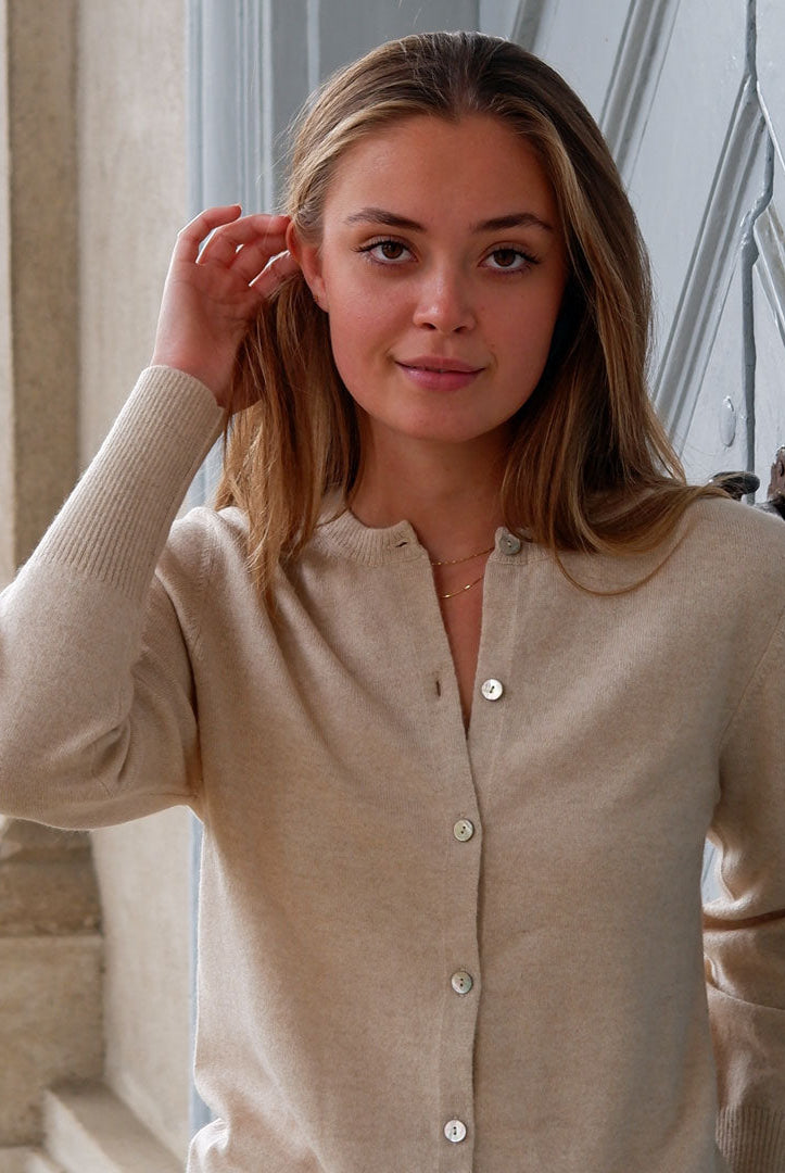 Kvinde iført en Soft Sand cashmere cardigan med knapper, står ved en grå dør og smiler.
