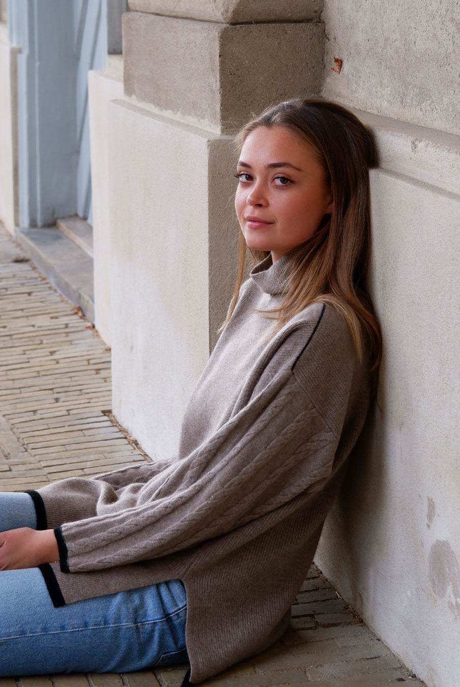 Model siddende ved sandfarvet væg iført Tuya cashmere mørk gråbeige rullekravebluse med mønster.