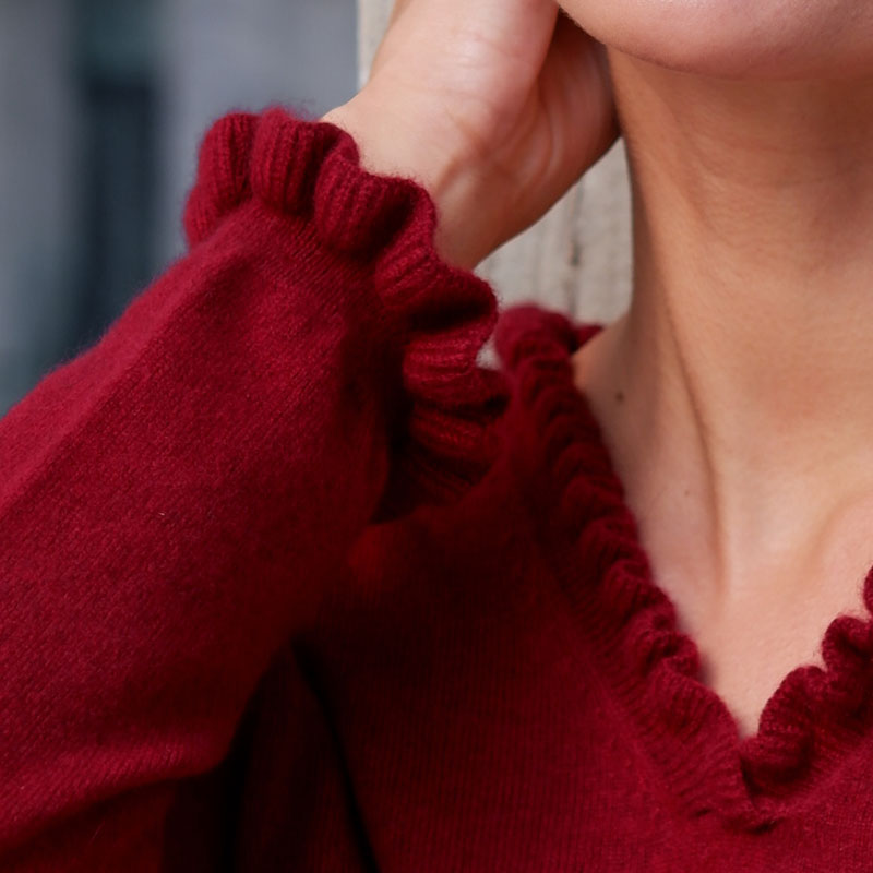 Nærbillede af flæsedetaljer ved halsudskæring og manchet på cashmere bluse i Pinotrød.