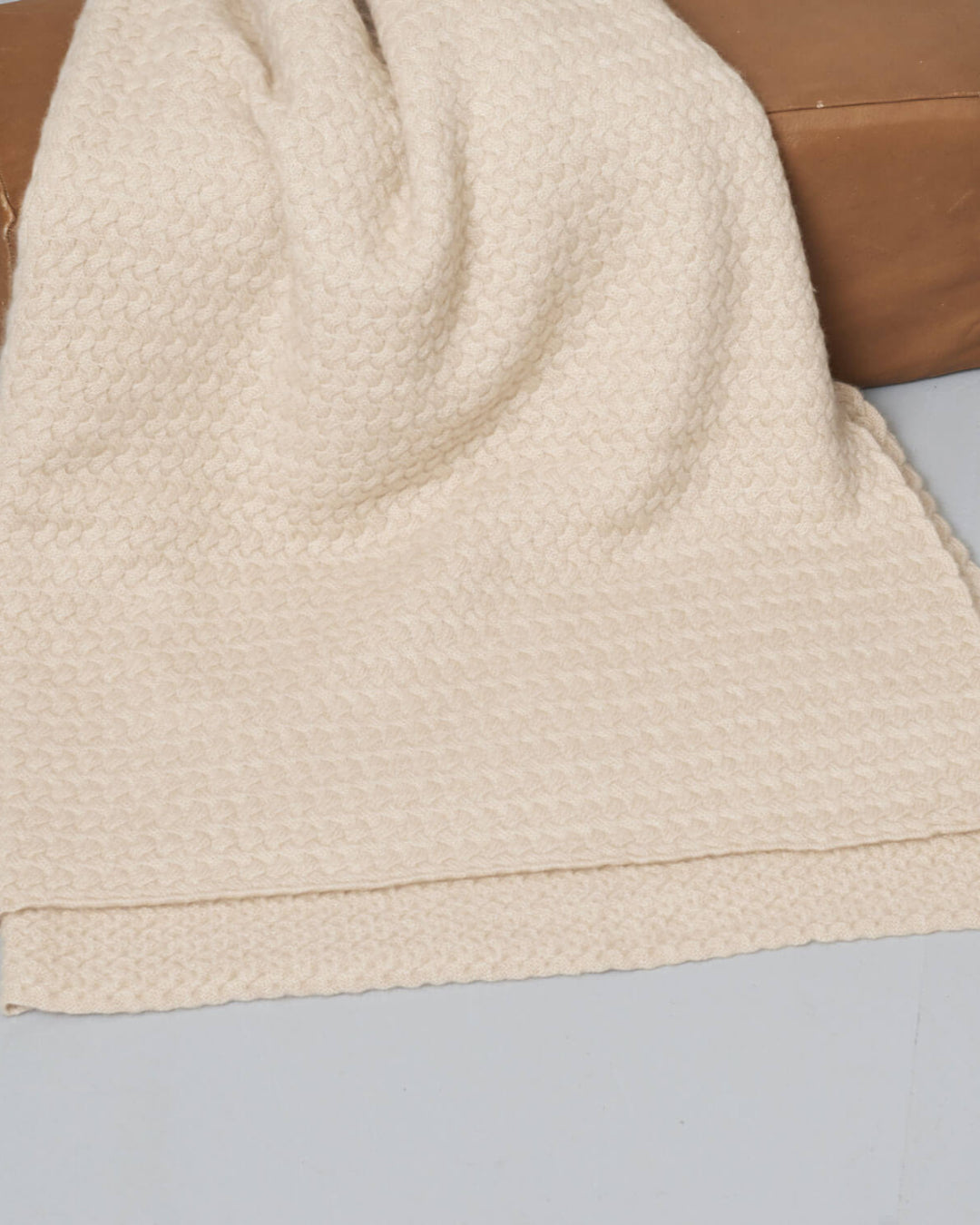 Plaiden i lys beige draperet over en læderpuf, der fremhæver den luksuriøse cashmere-kvalitet og det tidløse design.