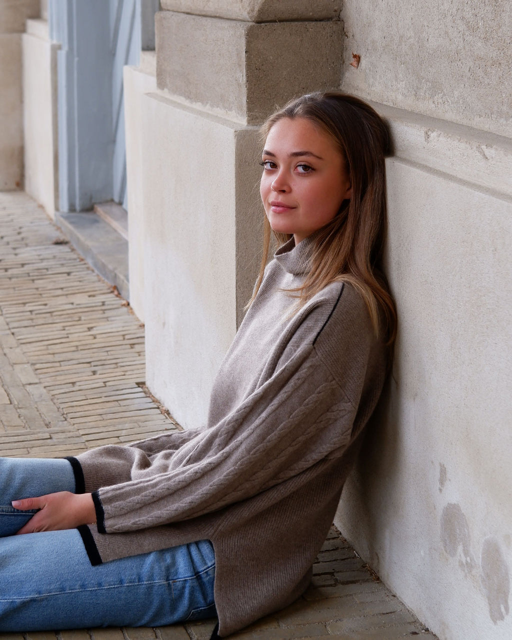 Model siddende ved sandfarvet væg iført Tuya cashmere mørk gråbeige rullekravebluse med mønster.