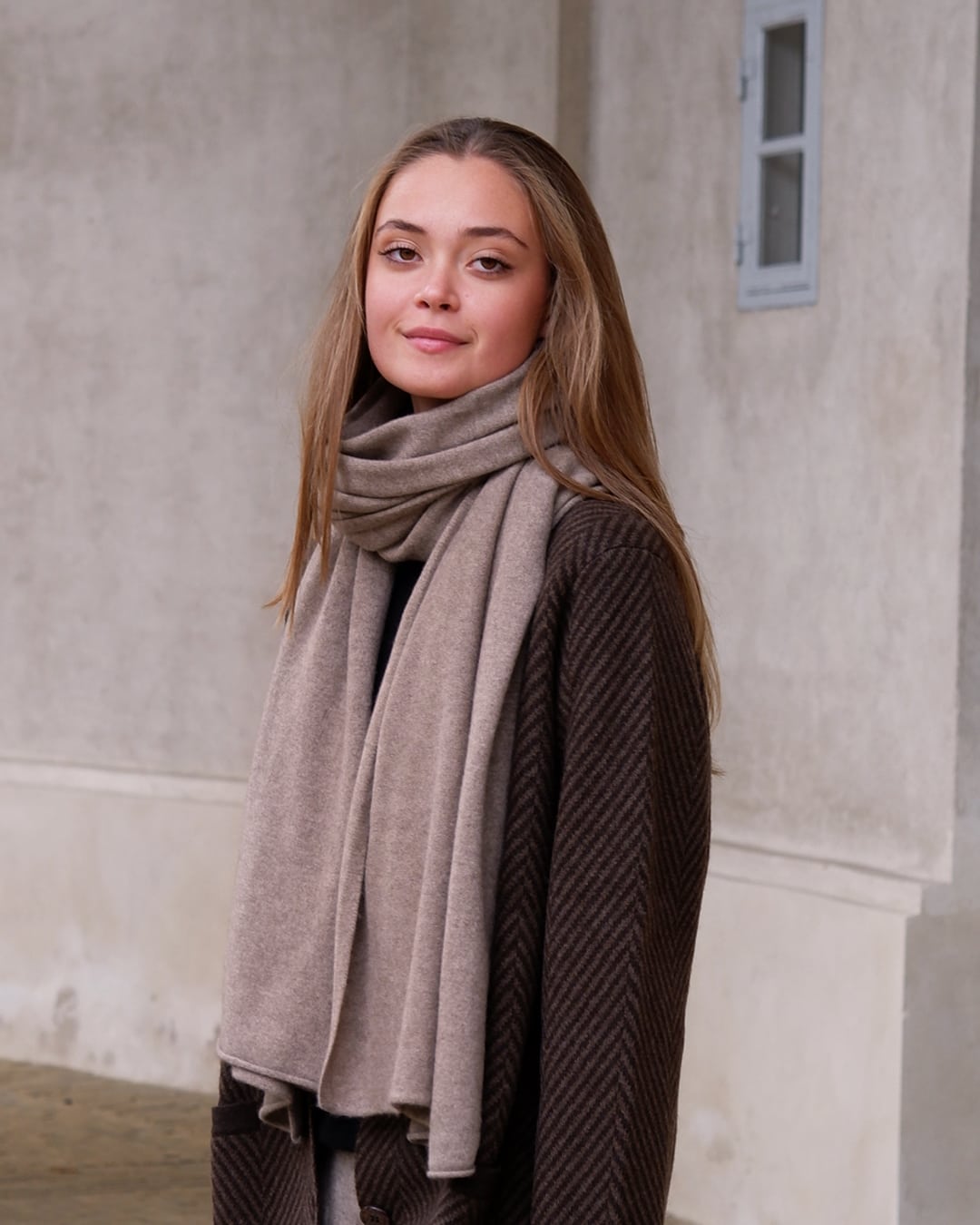 Model med det store naturfarvede cashmere tørklæde i mørk gråbeige svøbt om halsen.